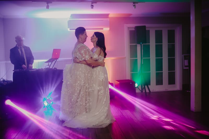 Two brides in lace wedding dresses embrace and dance on a wooden floor with colorful stage lights and a DJ in the background.