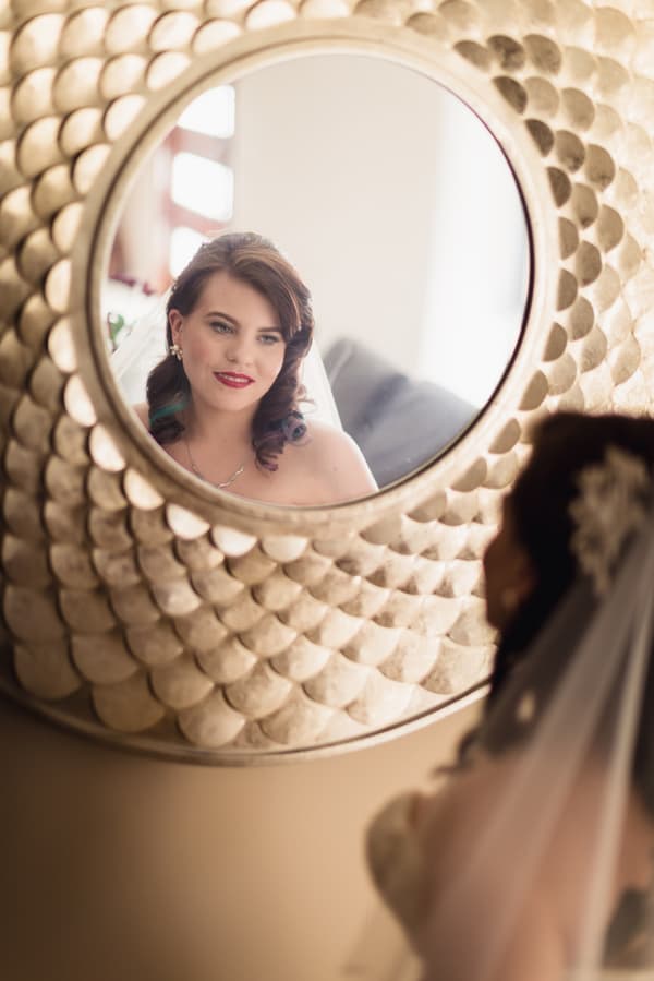 The bride Holly is reflected in a decorative round mirror as she looks at herself, wearing her wedding veil and earrings at Kwila Lodge.