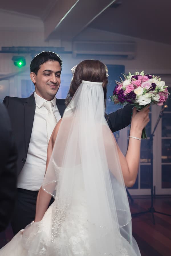 The groom smiles at the bride who is holding a bouquet of flowers at Hillstone St Lucia — The Rosewood Room during the reception.
