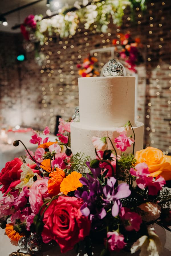 Two-tier white wedding cake decorated with small disco balls, surrounded by a vibrant floral arrangement featuring roses and other colorful flowers at Sandstone Point Hotel — Cellar.