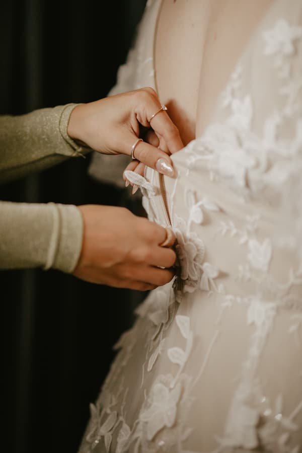 Close-up of hands fastening the back zipper of a floral lace wedding dress.