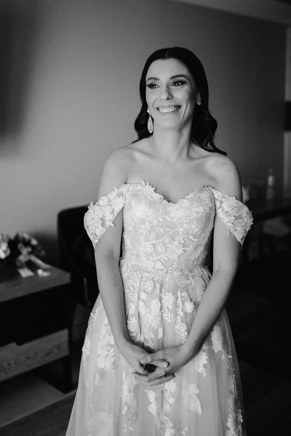 Bride in an off-shoulder lace wedding dress standing indoors, smiling and looking slightly to her left.