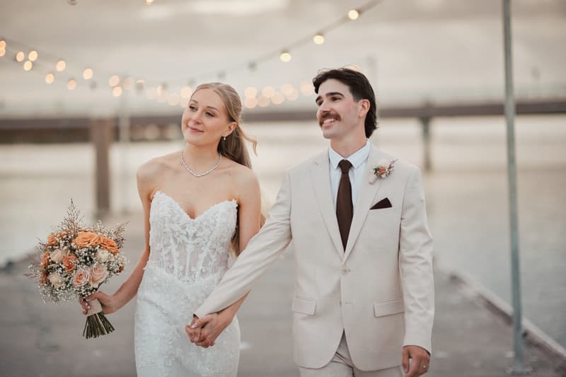 Morgan and Joel hold hands and walk outdoors near water at Sandstone Point Hotel during their couple portraits session. Morgan wears a strapless lace wedding gown and holds a bouquet of peach and cream roses, while Joel wears a light beige suit with a dark tie and boutonniere.