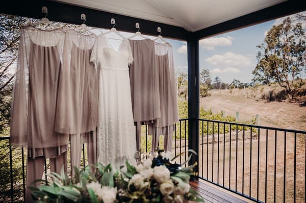 The bride's white lace wedding dress and four bridesmaids' taupe dresses hang on hangers from a covered porch railing, with a floral arrangement in the foreground and a rural landscape in the background. Wedding photographers Brisbane by She Said Yes.