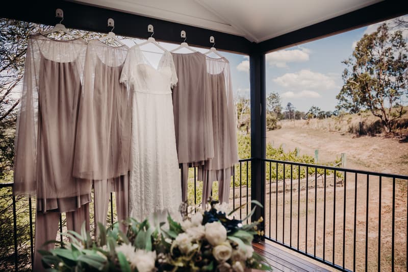 The bride's white lace wedding dress and four bridesmaids' taupe dresses hang on hangers from a covered porch railing at Ocean View Estates, with a floral arrangement in the foreground and a rural landscape in the background.