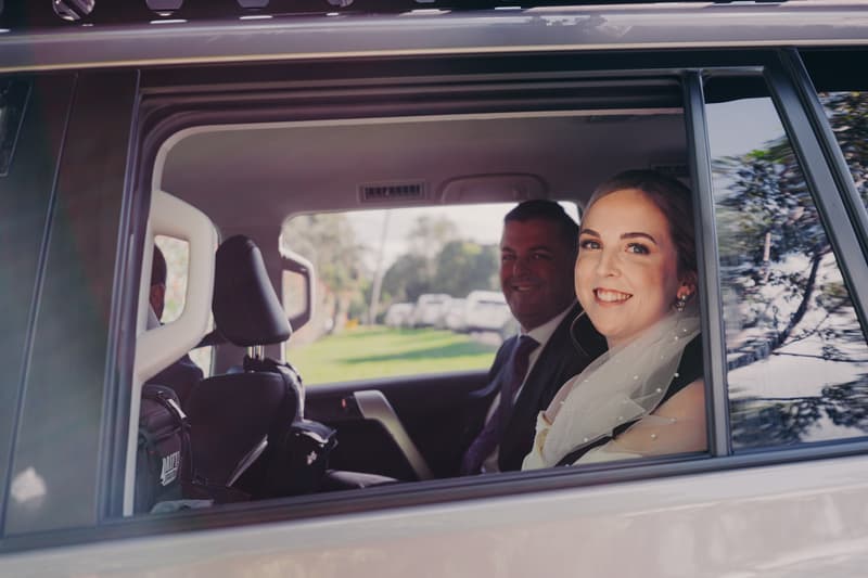 Courtney the bride and Liam the groom sit inside a car, visible through the side window, with Courtney smiling at the camera.