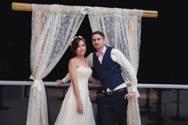 The bride Wing and groom Jason pose together on the reception stage at Bilinga Beach Weddings — Bilinga SLSC, standing in front of a decorative lace curtain backdrop at night.