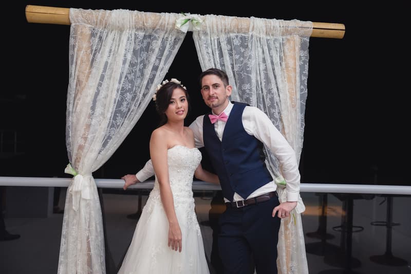 The bride Wing and groom Jason pose together on the reception stage at Bilinga Beach Weddings — Bilinga SLSC, standing in front of a decorative lace curtain backdrop at night.