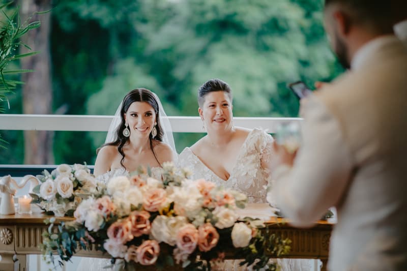 Two brides in wedding dresses sit behind a table adorned with a large floral arrangement, while a man in a light-colored suit takes their photo with a smartphone.
