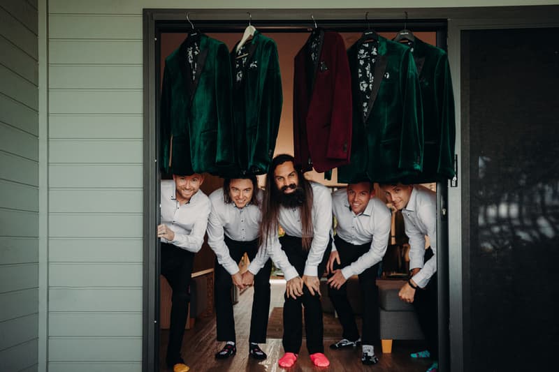 Five groomsmen crouch and smile under hanging green and burgundy velvet jackets at Sandstone Point Hotel.