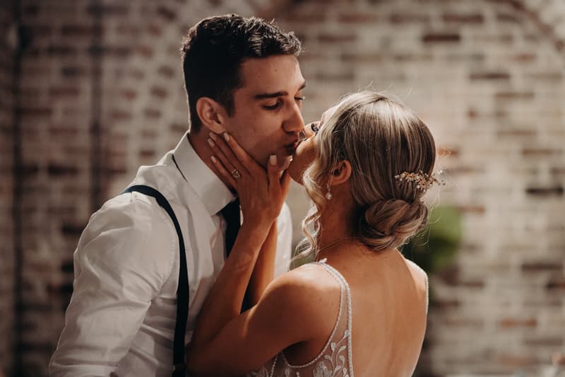 Courtney and Cameron share a kiss at the reception stage of Sandstone Point Hotel — Cellar, with Courtney wearing a detailed bridal gown and Cameron in a white shirt with suspenders.