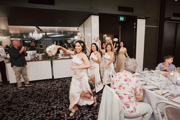 Bridesmaids in matching satin dresses hold bouquets and walk in a line at the reception in The Pumicestone Room,, while guests seated at tables watch and applaud. What is an engagement party by She Said Yes.