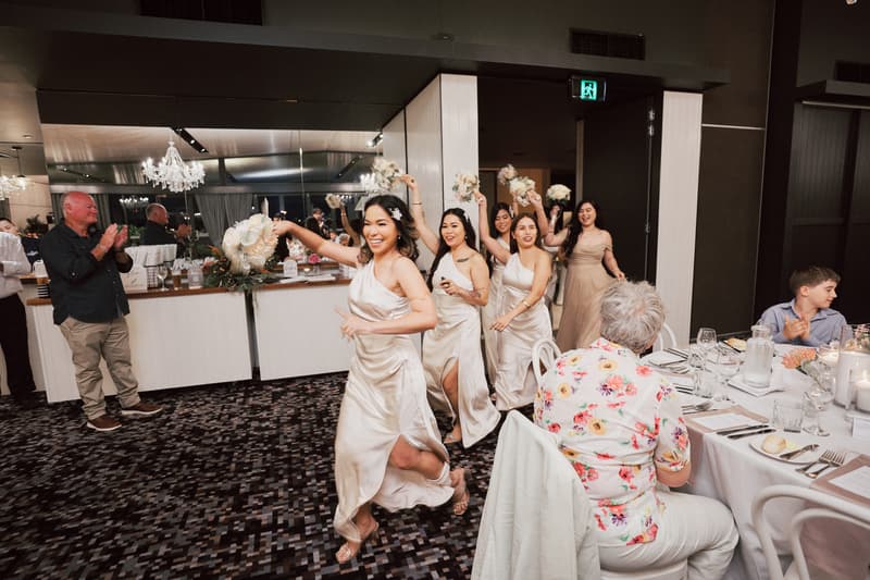 Bridesmaids in matching satin dresses hold bouquets and walk in a line at the reception in The Pumicestone Room, Sandstone Point Hotel, while guests seated at tables watch and applaud.