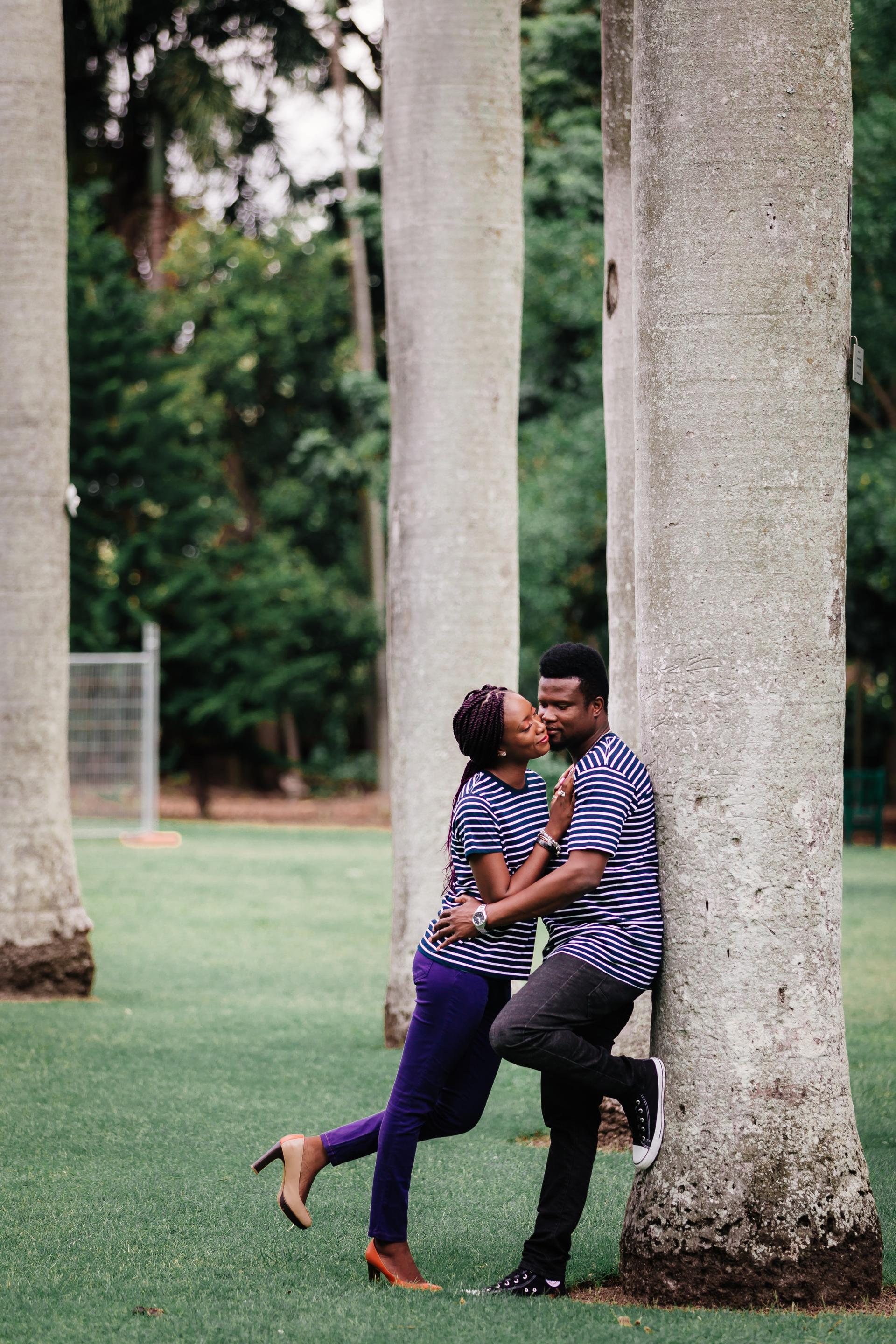 The couple shares a kiss while leaning against a tree in Brisbane's City Botanic Gardens, surrounded by lush greenery.
