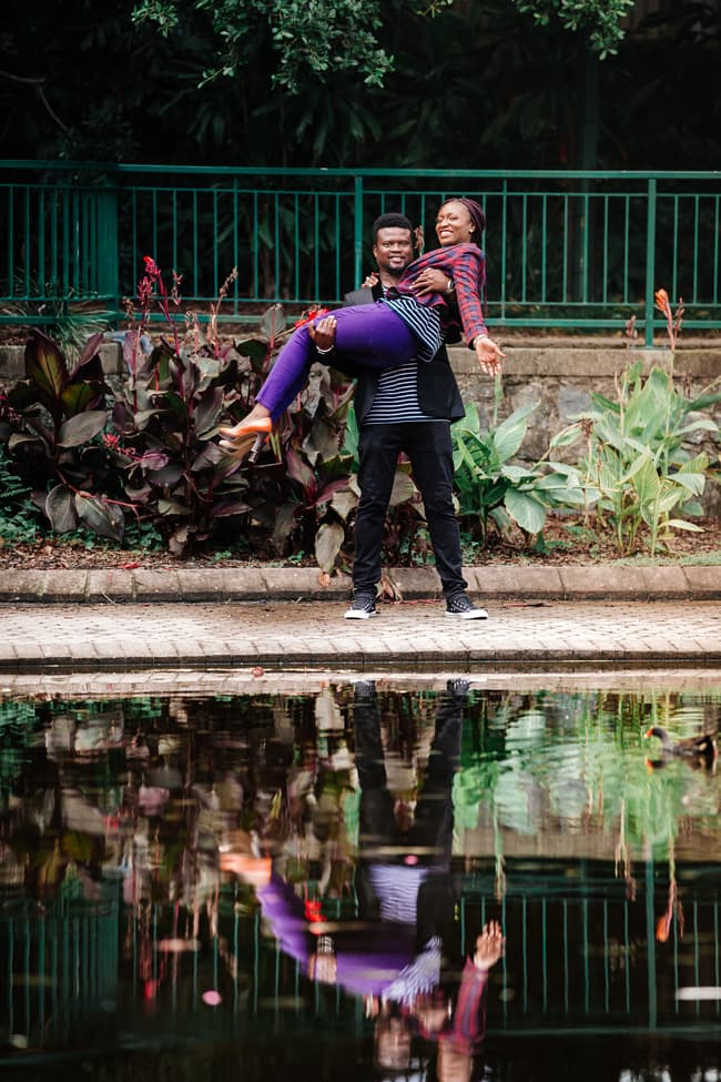The couple playfully poses by a pond, with one lifting the other, surrounded by vibrant plants in Brisbane's City Botanic Gardens.