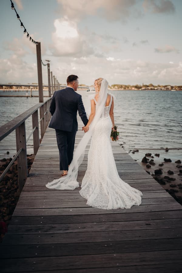 The bride Krystal and groom Brandon walk hand in hand along a wooden pier at Sandstone Point Hotel, with the bride holding a bouquet and wearing a long veil and lace gown.