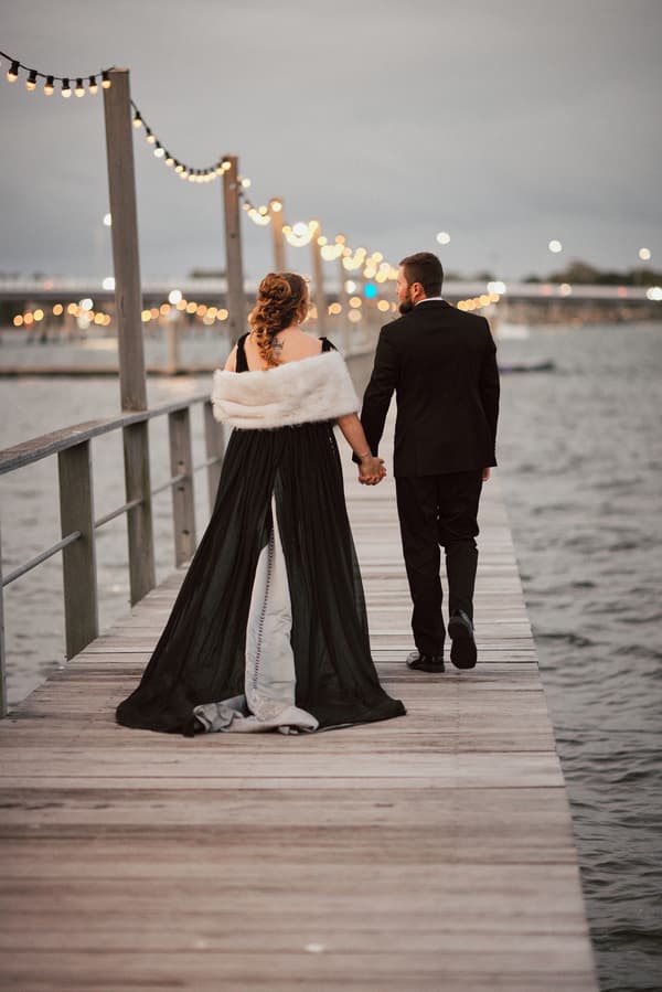 The bride Jayme and groom Christopher walk hand in hand along a wooden pier decorated with string lights over water at Sandstone Point Hotel.