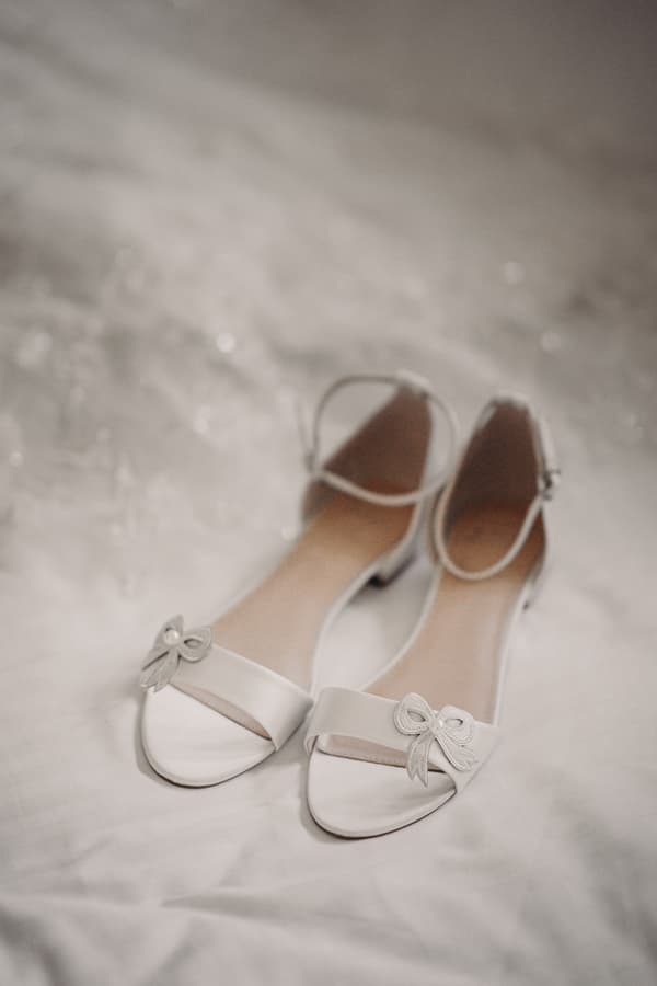 Close-up of the bride's white open-toe shoes with ankle straps and decorative bows, placed on a white fabric surface.