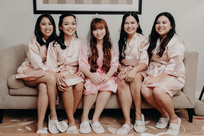 The bride and four bridesmaids sit on a couch at Royal on the Park wearing matching satin robes and slippers labeled with their roles.