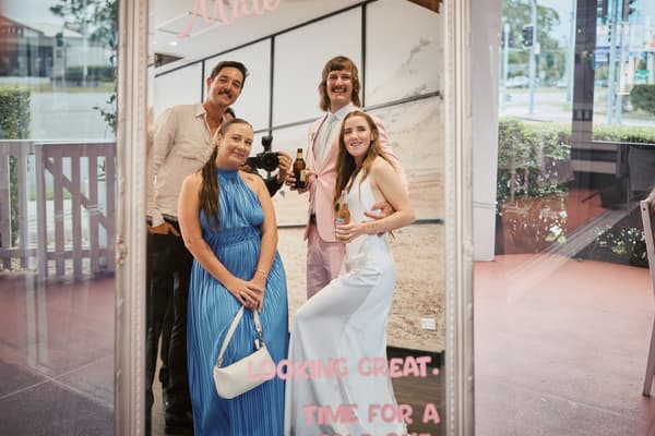 The bride Olivia in a white dress and the groom Jake in a pink suit pose with two guests in front of a large mirror at White Horse Ranch during the reception stage. Olivia holds a drink, Jake holds a beer bottle, and one guest holds a camera.