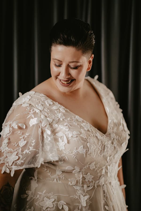 A bride wearing a white lace wedding dress with floral embroidery stands against a dark curtain background, looking down and smiling.