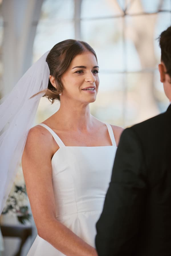 The bride Ashleigh in a white wedding dress and veil faces the groom James during their ceremony at Sandstone Point Hotel — The Pavilion.