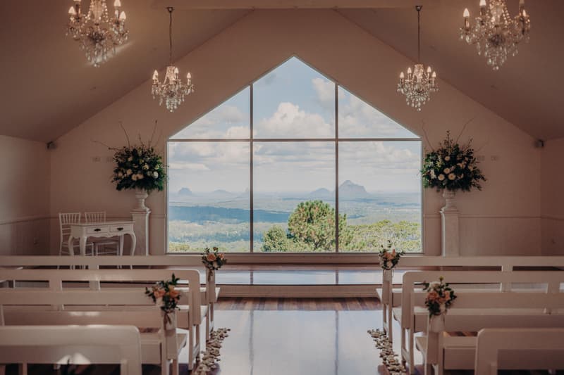 Empty ceremony chapel at Tiffany's Maleny with white benches decorated with floral arrangements, crystal chandeliers, and a large window showing a scenic mountain view.