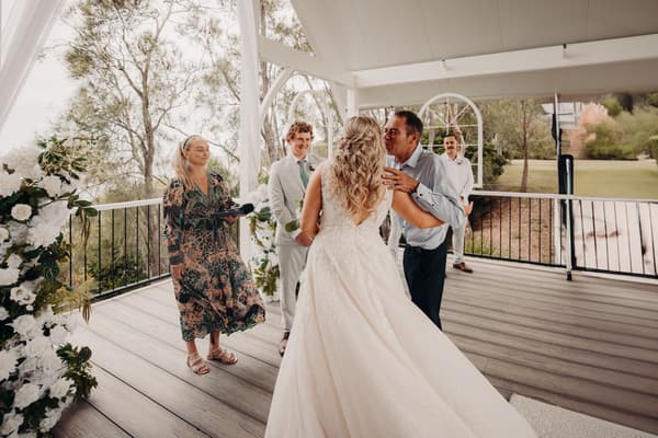 The bride Chloe is kissed on the cheek by an older man, likely her father, at the Sandstone Point Hotel — Pavilion during the ceremony. The groom Brodie stands nearby in a light-colored suit, smiling. A female celebrant holds a microphone and a man in white stands in the background. The pavilion is decorated with white flowers and greenery.