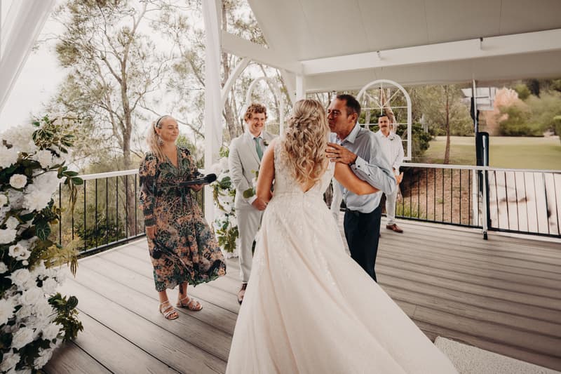 The bride Chloe is kissed on the cheek by an older man, likely her father, at the Sandstone Point Hotel — Pavilion during the ceremony. The groom Brodie stands nearby in a light-colored suit, smiling. A female celebrant holds a microphone and a man in white stands in the background. The pavilion is decorated with white flowers and greenery.