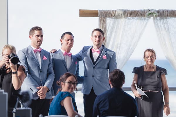 Jason and two groomsmen stand at the ceremony stage at Bilinga Beach Weddings — The Terrace, with the officiant holding papers nearby and guests seated in front.