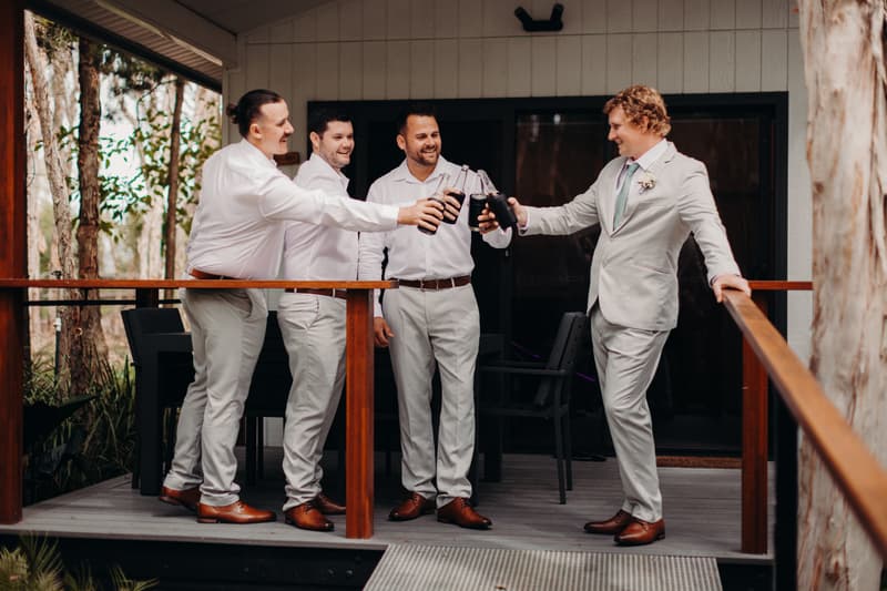 Brodie and three groomsmen toast with bottles on a wooden deck at Sandstone Point Hotel.