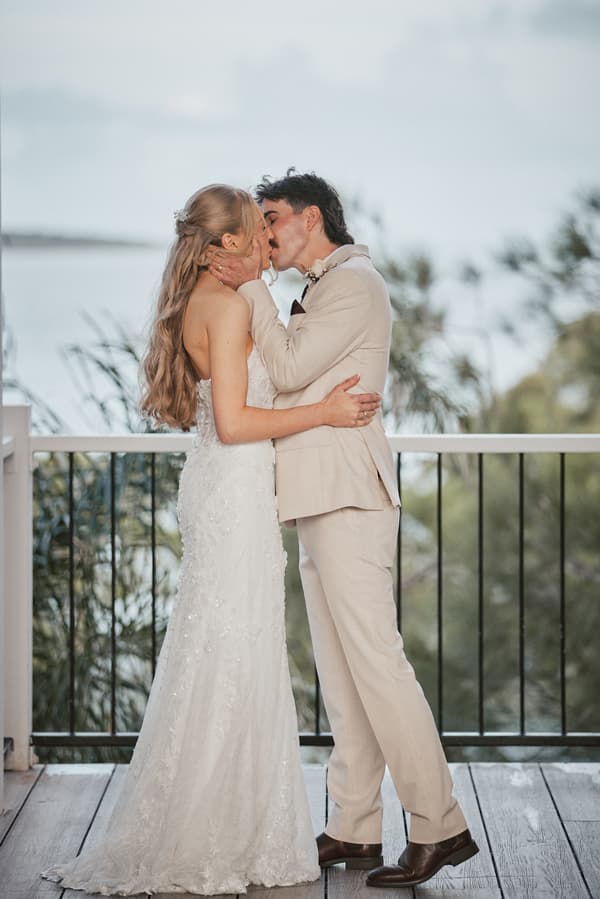 Morgan and Joel share a kiss on the ceremony stage at Sandstone Point Hotel — The Pavilion, with a natural outdoor backdrop visible behind the railing.