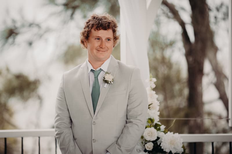 The groom Brodie stands at the Pavilion of Sandstone Point Hotel during the ceremony, wearing a light grey suit with a green tie and a white boutonniere.