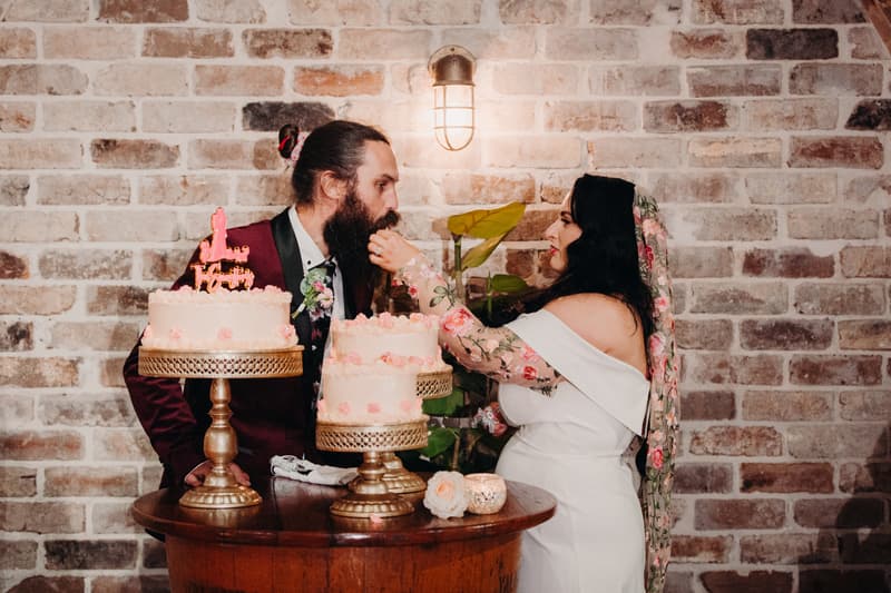 The bride Mindy feeds cake to the groom David at a small round wooden table with two tiered cakes on gold stands at Sandstone Point Hotel — Cellar.