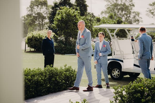 Three men in light blue suits with pink boutonnieres stand near a white golf cart outside, while a woman in dark clothing stands nearby on the grass. Getting ready photos by She Said Yes.