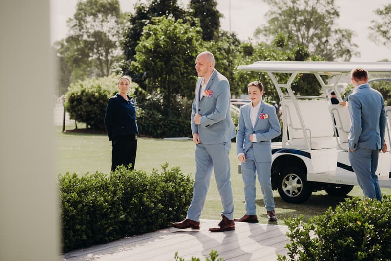 Three men in light blue suits with pink boutonnieres stand near a white golf cart outside at Sandstone Point Hotel — Pavilion, while a woman in dark clothing stands nearby on the grass.