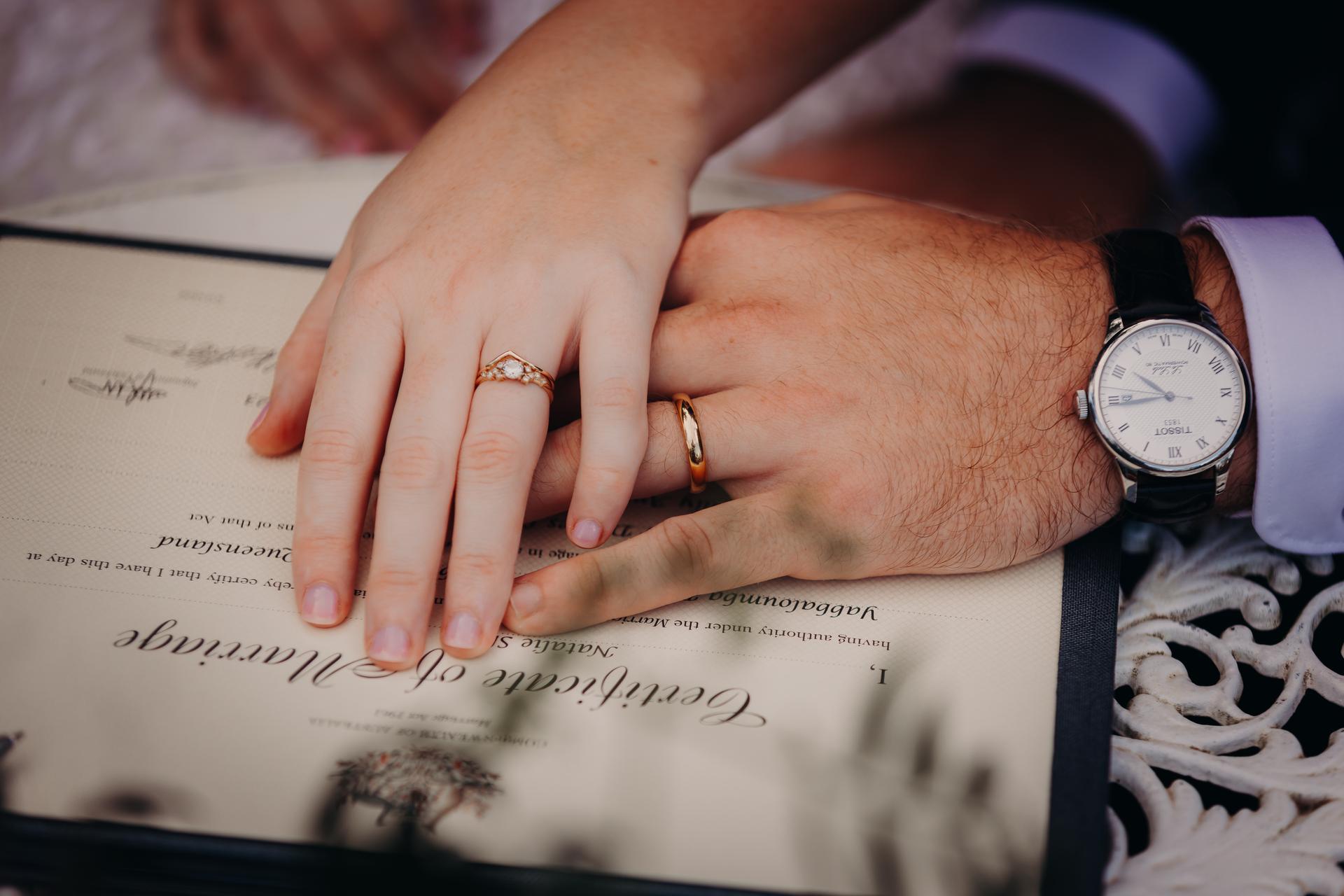 The bride and groom place their hands on a signed marriage certificate at Yabbaloumba Retreat — By The River, showing their wedding rings.