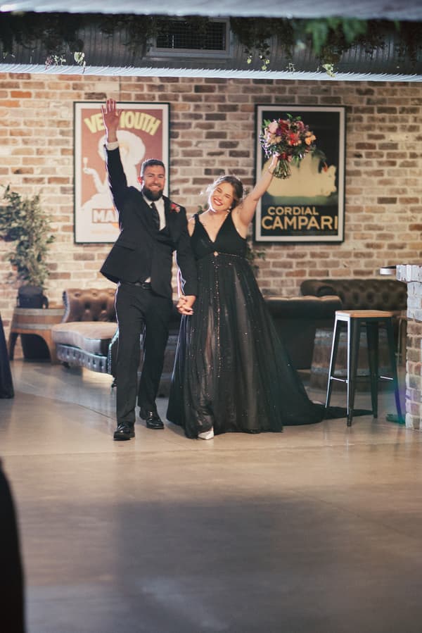 Bride Jayme in a black gown and groom Christopher in a black suit hold hands and raise their arms while entering the reception at Sandstone Point Hotel — The Cellar.