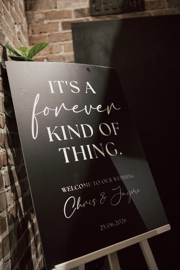 Wedding welcome sign at Sandstone Point Hotel — The Cellar reading 'It's a forever kind of thing. Welcome to our wedding Chris & Jayme 25.06.2026' displayed on an easel against a brick wall.