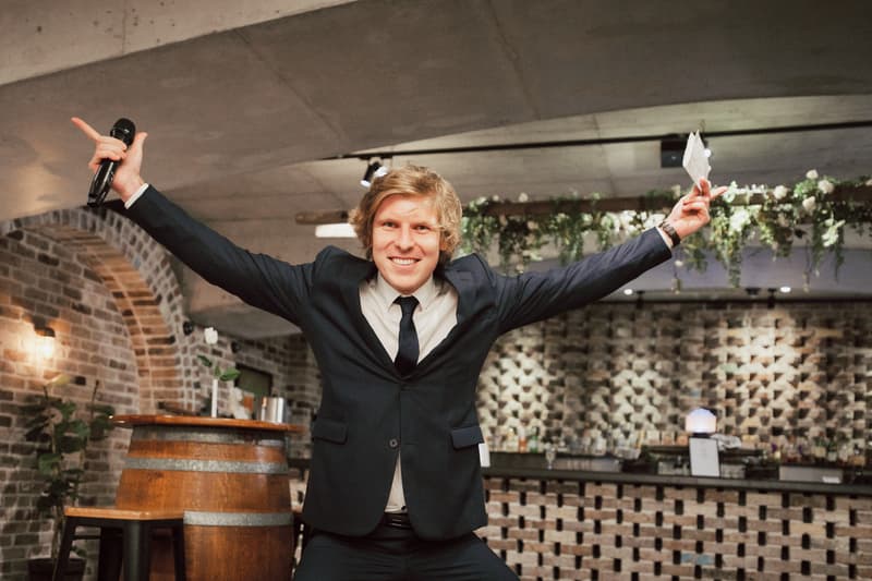 A man in a dark suit and tie holds a microphone and a piece of paper with arms outstretched inside the Sandstone Point Hotel — The Cellar reception area with brick walls and a wooden barrel table.