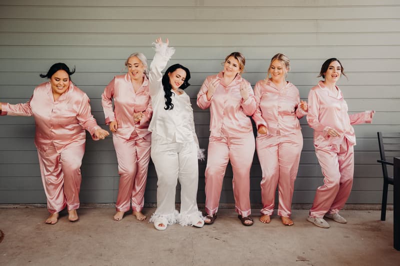 The bride and five bridesmaids wearing matching satin pajamas pose and dance against a gray wooden wall at Sandstone Point Hotel — Pavilion.