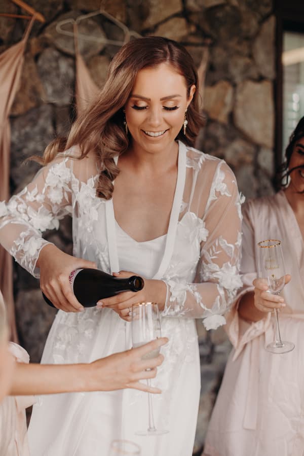 The bride Chelsea, wearing a white floral lace robe, pours champagne into a glass held by a bridesmaid at Preston Peak Winery Functions Centre.
