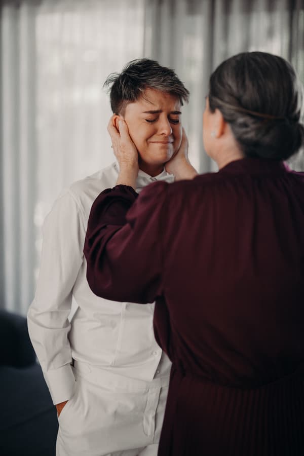 Tiffany in a white shirt and pants has her eyes closed while an older woman, possibly her mother, holds her face gently indoors at Sandstone Point Hotel — The Pavilion.