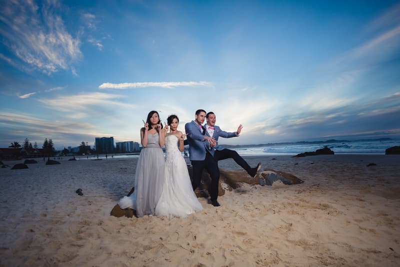The bride Wing, groom Jason, a bridesmaid, and a groomsman pose playfully on the beach at Bilinga Beach Weddings during couple portraits at sunset.