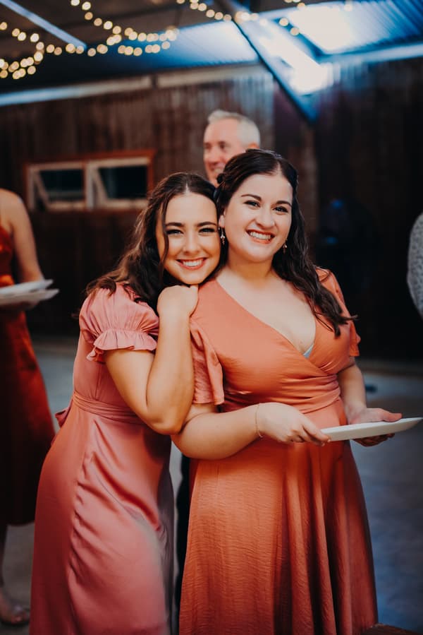 Two bridesmaids smile and pose together at the reception in The Shed at Yabbaloumba Retreat, one holding a plate.