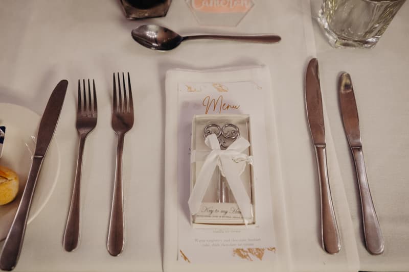 Close-up of a wedding reception table setting at Sandstone Point Hotel — Pumicestone Room featuring a menu card with a decorative key tied with a white ribbon, surrounded by cutlery, a glass, and a bread roll on a plate.