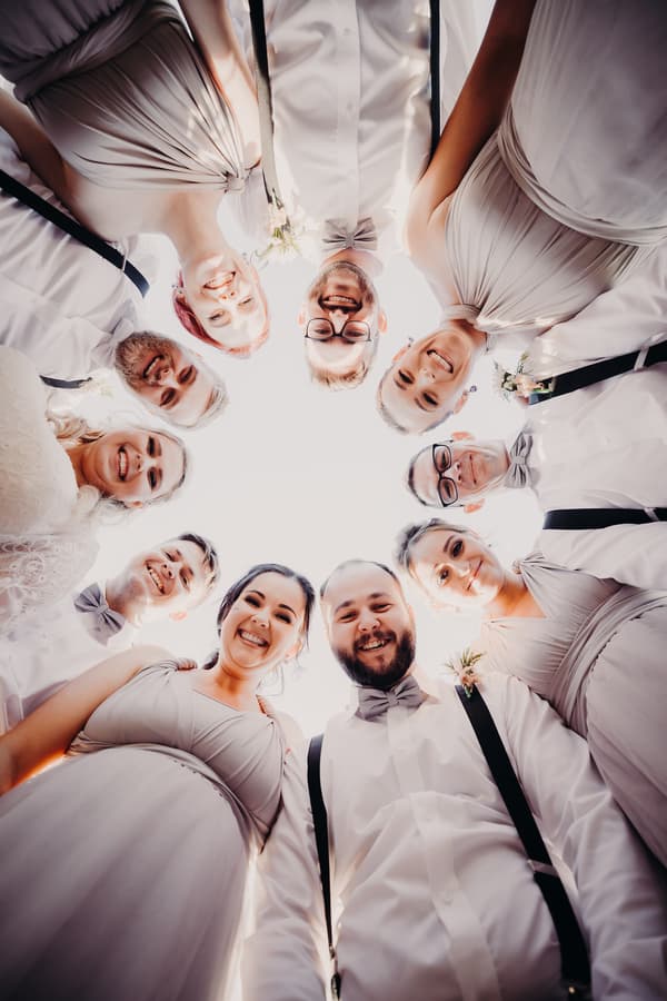 The wedding party, including the bride and groom, stand in a circle looking down at the camera, dressed in formal attire with the bridesmaids in matching dresses and groomsmen in white shirts with suspenders and bow ties.