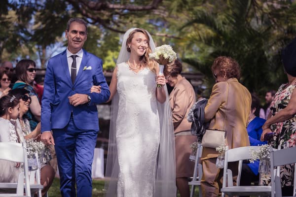 The bride Francesca, holding a bouquet, walks arm-in-arm with an older man, likely her father, down the aisle at TOOWONG ROWING CLUB — Brisbane River's Edge, while guests seated on white chairs watch and smile.