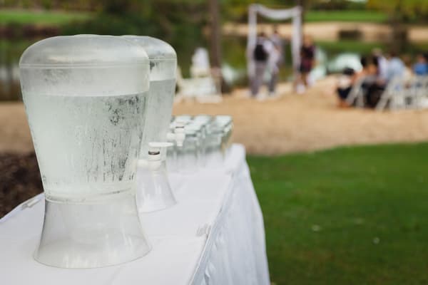 Two large water dispensers and a row of glasses on a white tablecloth-covered table outdoors at Novotel Sunshine Coast Resort, with blurred guests seated and standing near a ceremony arch in the background.