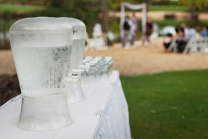 Two large water dispensers and a row of glasses on a white tablecloth-covered table outdoors at Novotel Sunshine Coast Resort, with blurred guests seated and standing near a ceremony arch in the background.